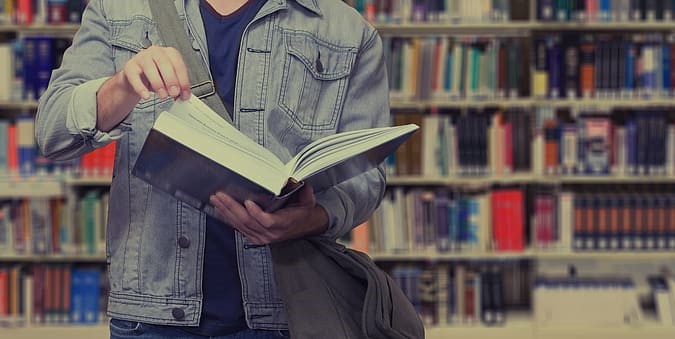 Na zdjęciu student z książką na tle półek z książkami